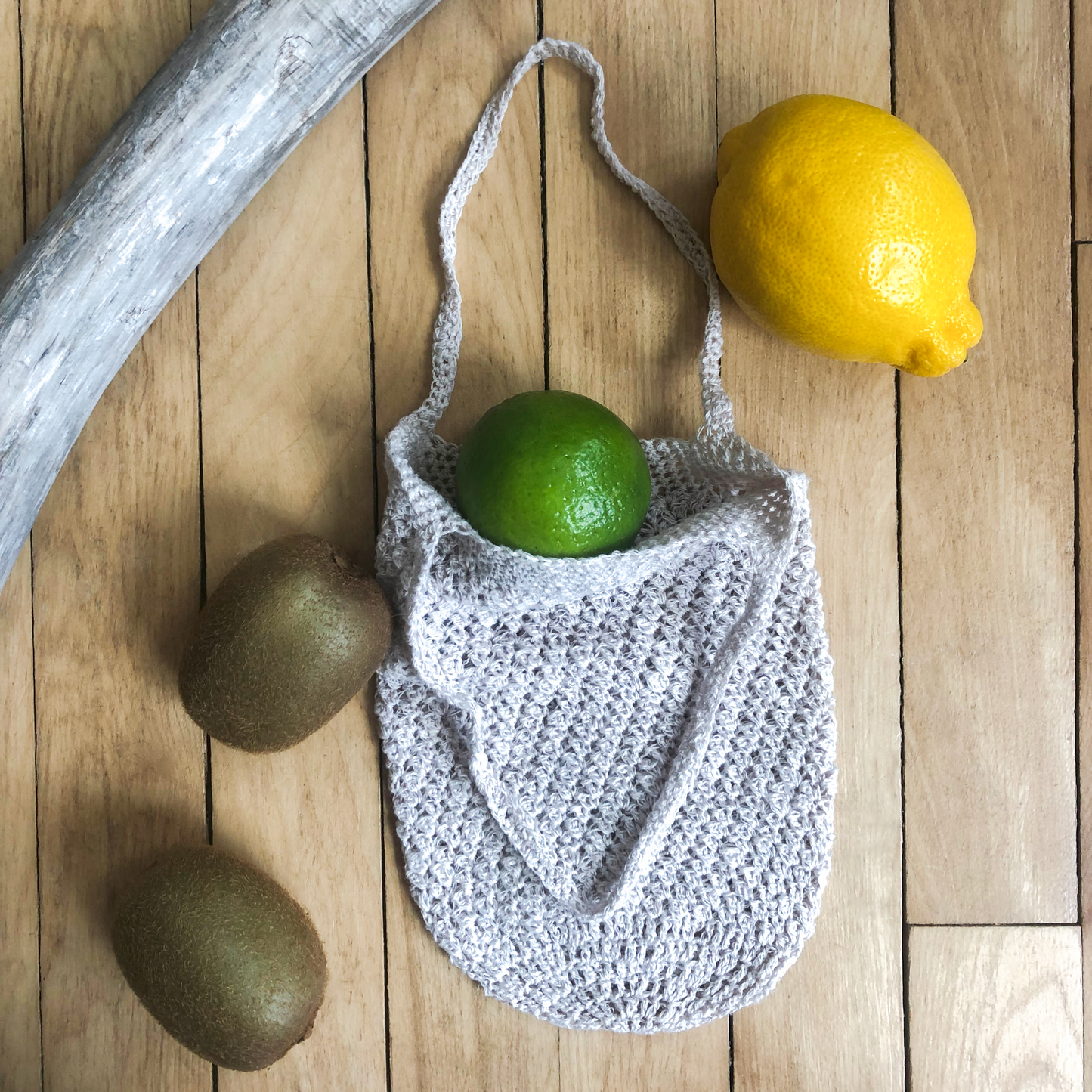 Small produce bag partially open with a lime coming out the top, two limes, a lemon, and a piece of driftwood laying beside the bag.