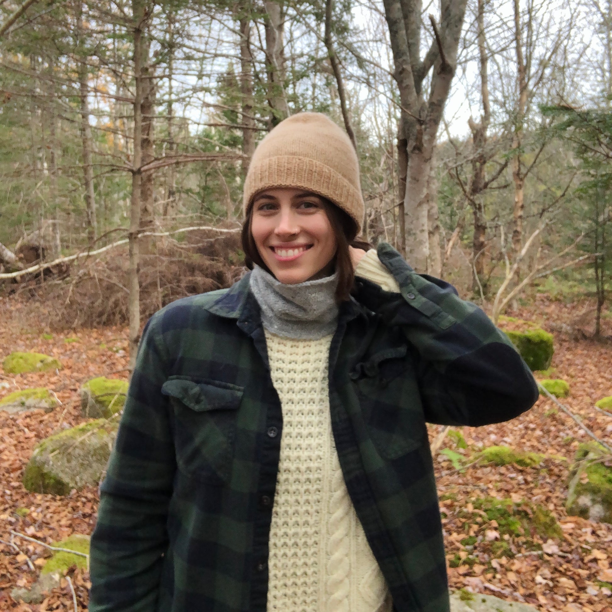 Pam wearing the grey nor'easter paired with the cashmere toque, dark green plaid shirt and cream knit sweater underneath. She is standing in the forest and posing for the camera.