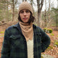 Pam modelling the cashmere bandana paired with matching cashmere toque, a dark green plaid shacket, and white knitted sweater underneath. The upcycled neck scarf drapes beautifully and falls in a short triangle against her chest.
