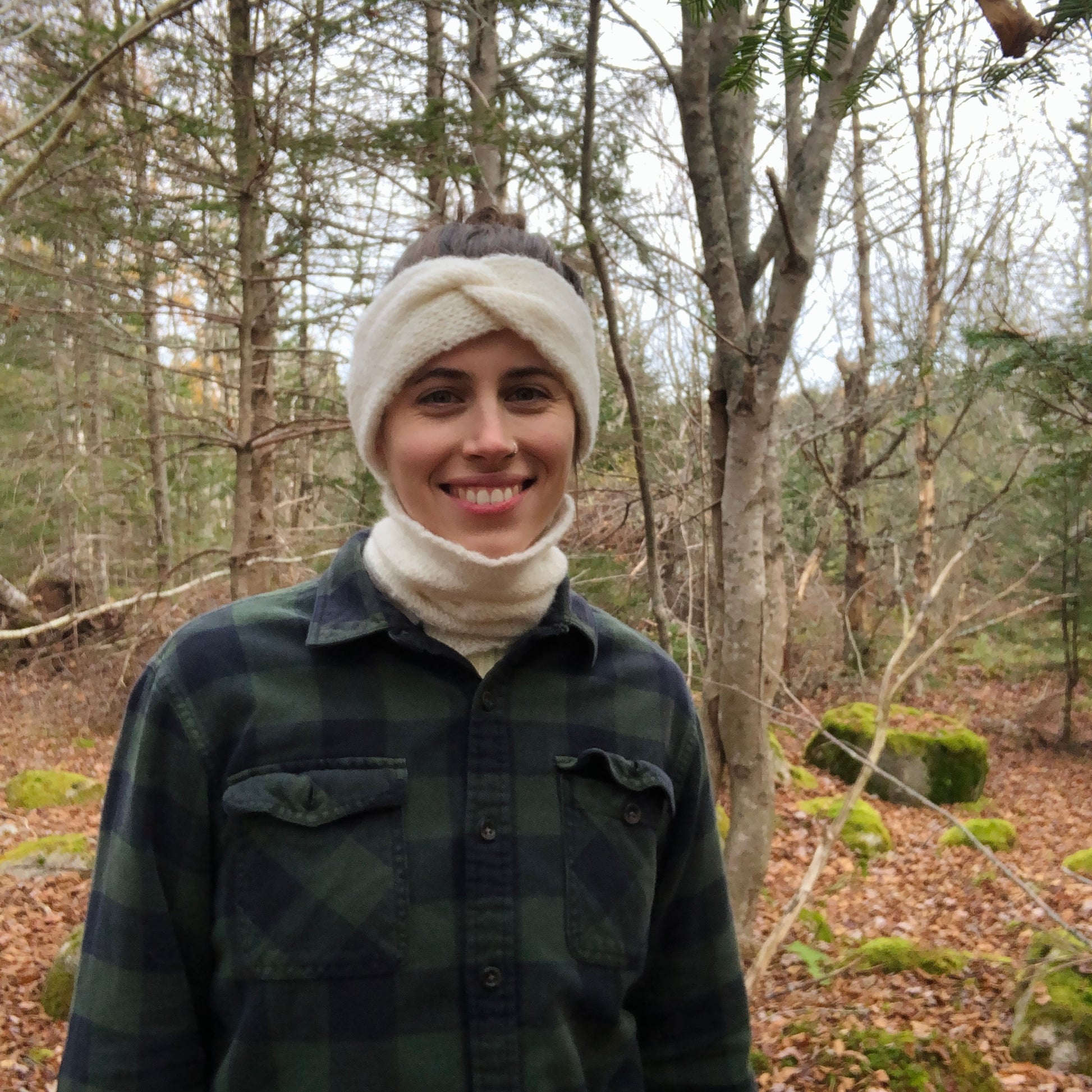 Pam is wearing the white audrey with the white wool neckwarmer as well. The two white and fuzzy wool options match perfectly together. She is also wearing a dark green plaid shirt and is smiling at the camera in the woods. 