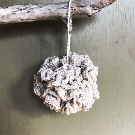 A crochet bath pouf hangs from a piece of driftwood in front of a muted green backdrop. The off-white cotton is crocheted so that many ripples form outward from the middle making a big squishy bath pouf!
