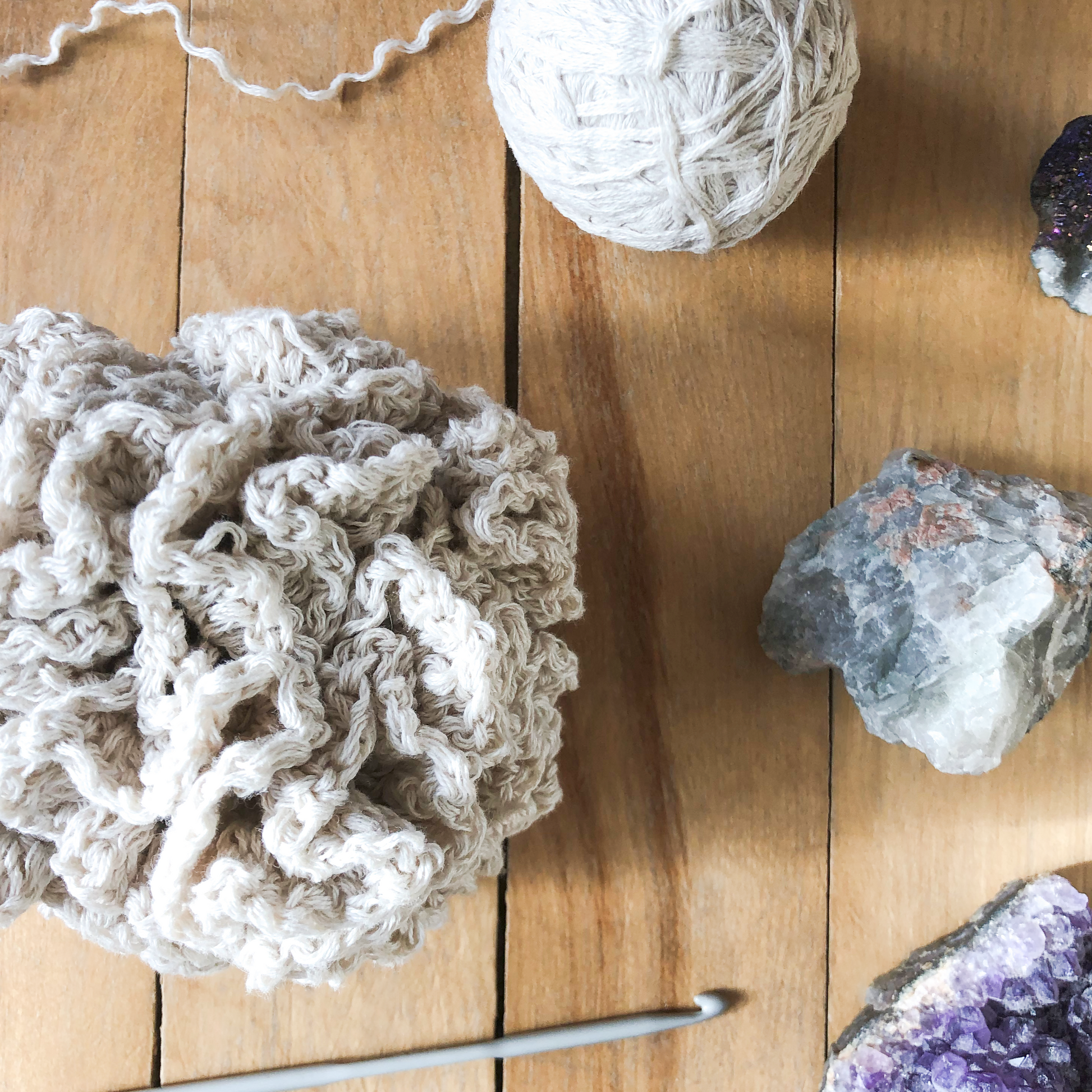 A size large crochet bath pouf, a ball of white cotton yarn, and gemstones lay against a wooden background. A metal crochet hook is laying beside the finished piece.