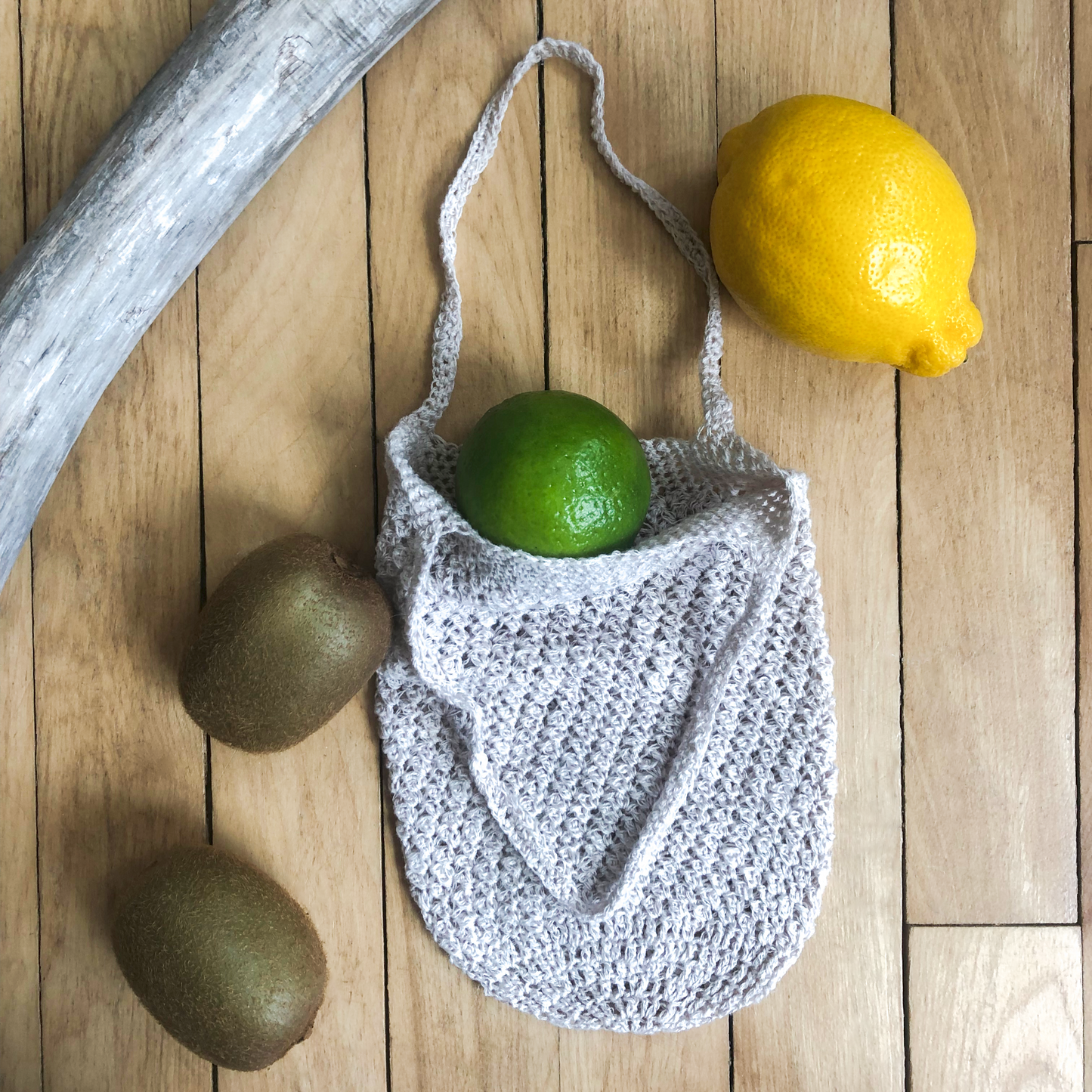 Small produce bag partially open with a lime coming out the top, two limes, a lemon, and a piece of driftwood laying beside the bag. 